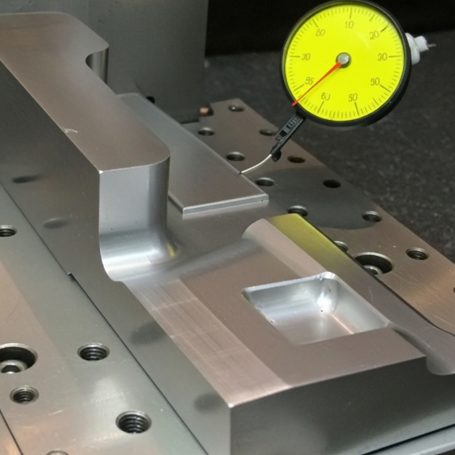 Pieza metálica con un dial de medición amarilla sobre una superficie de trabajo.